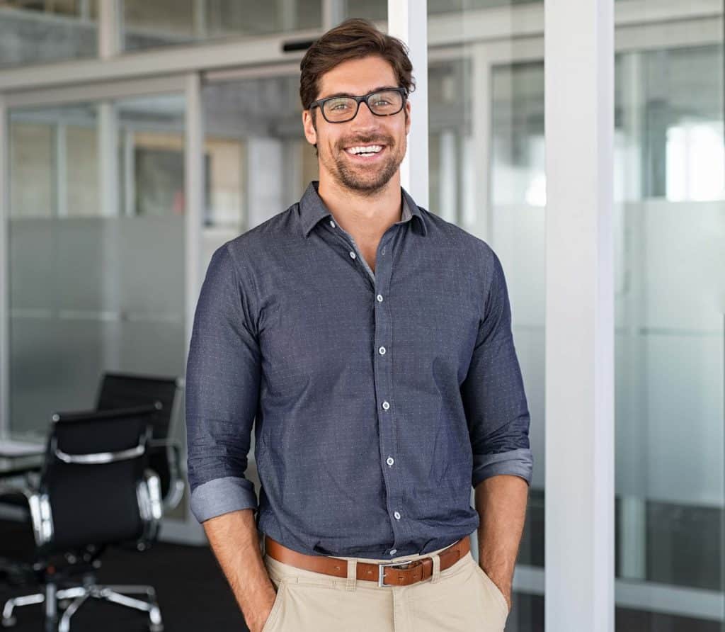 Good looking, smiling and confident, 30-something man in an office environment, wearing black-framed glasses, shortish dark brown hair parted on the side and feathered, with short facial hair, very white teeth, wearing a dark navy collared shirt with a light pattern, brown belt, and khakis with his hands in his pants pockets.
