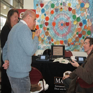 One man and one woman standing looking at and talking to one man that is seated at what appears to be a tradeshow booth/table
