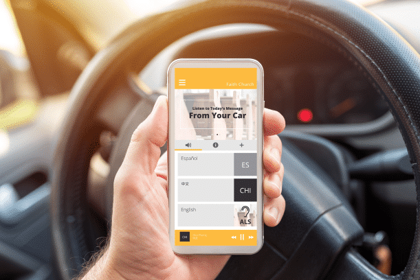 Man holding a phone, with a steering wheel of a car in the background. On the cell phone it shows the Listen EVERYWHERE app and channels to pick from.