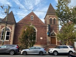 An older church building with red bricks and brown roofing. Three cars are parked out in front on the street.